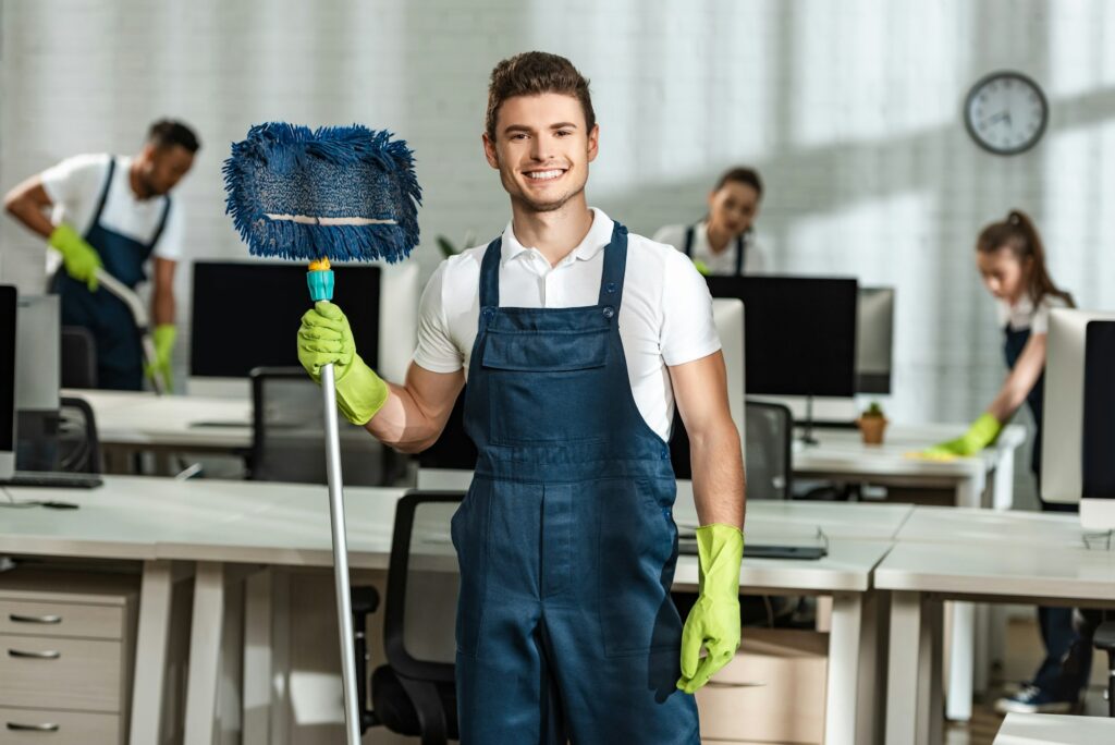 cheerful cleaner holding mop while looking at camera near multicultural colleagues