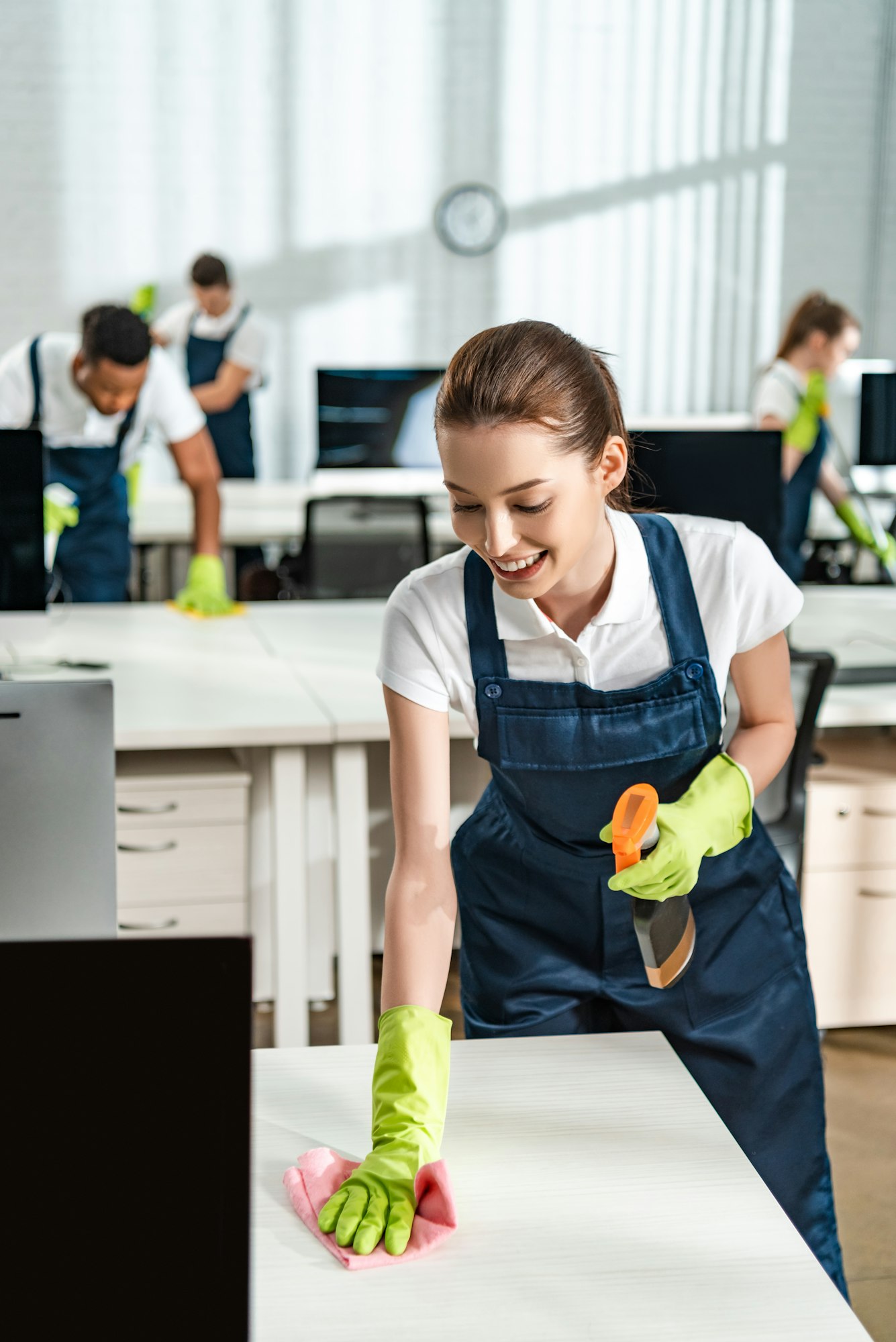 cheerful cleaner in overalls cleaning office desk with rag