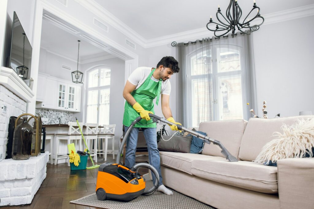 Indian male cleaner cleaning sofa with modern vacuum cleaner