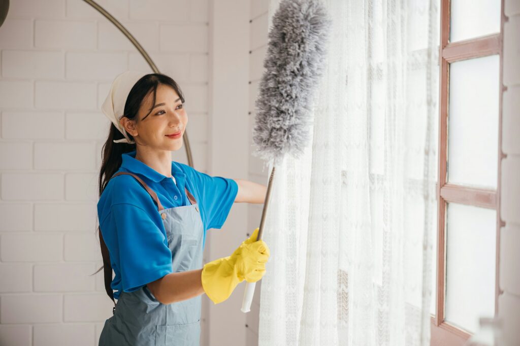 Enjoyment in routine cleaning for this smiling Asian woman. She stands dusting window blinds with