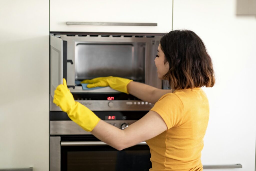 Happy Young Arab Woman Wiping Microwave With Rag While Cleaning In Kitchen