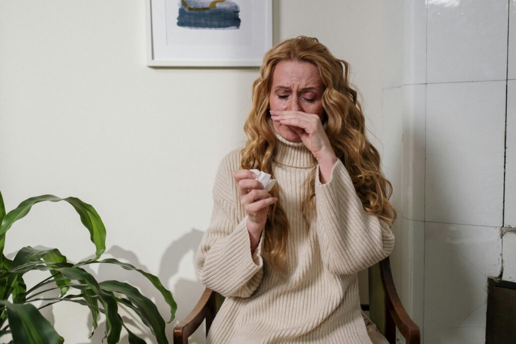 A woman with long, wavy blonde hair sits in a chair indoors, wearing a beige turtleneck sweater. She appears to be experiencing allergy symptoms or a cold, holding a tissue in one hand and touching her nose with the other. Her facial expression suggests discomfort, and there is a green houseplant in the foreground, adding a touch of nature to the scene. The background features a white wall with a framed painting and tiled sections.